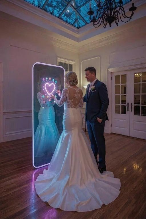 Bride and groom interact with a digital mirror displaying a heart symbol in an elegantly decorated room.